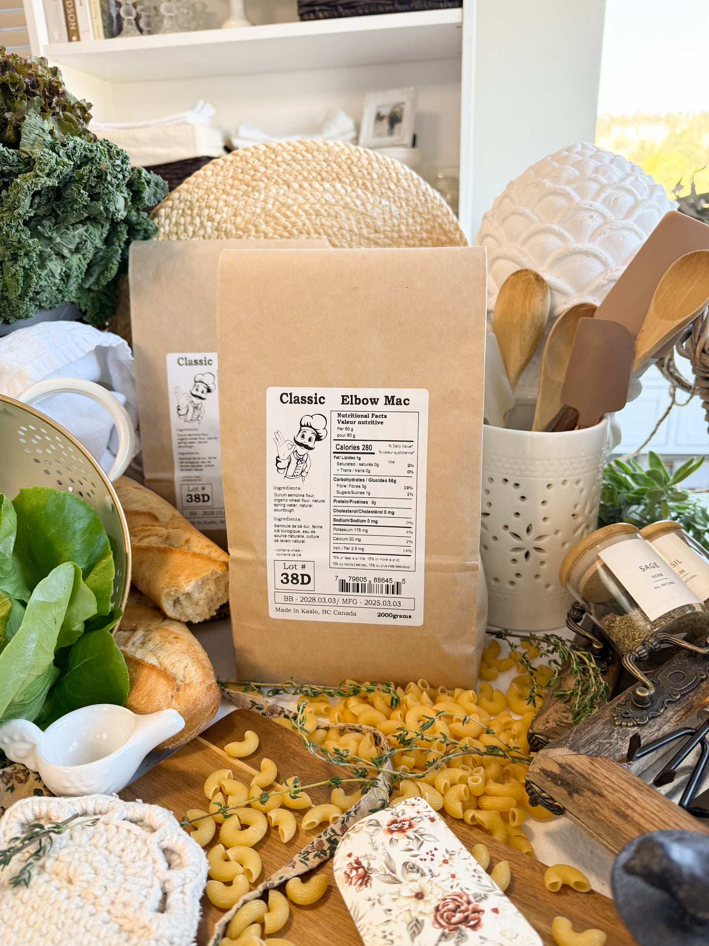 A kraft paper bag of Classic Elbow Mac pasta is displayed on a rustic wooden table surrounded by fresh ingredients, bread, and kitchen utensils.