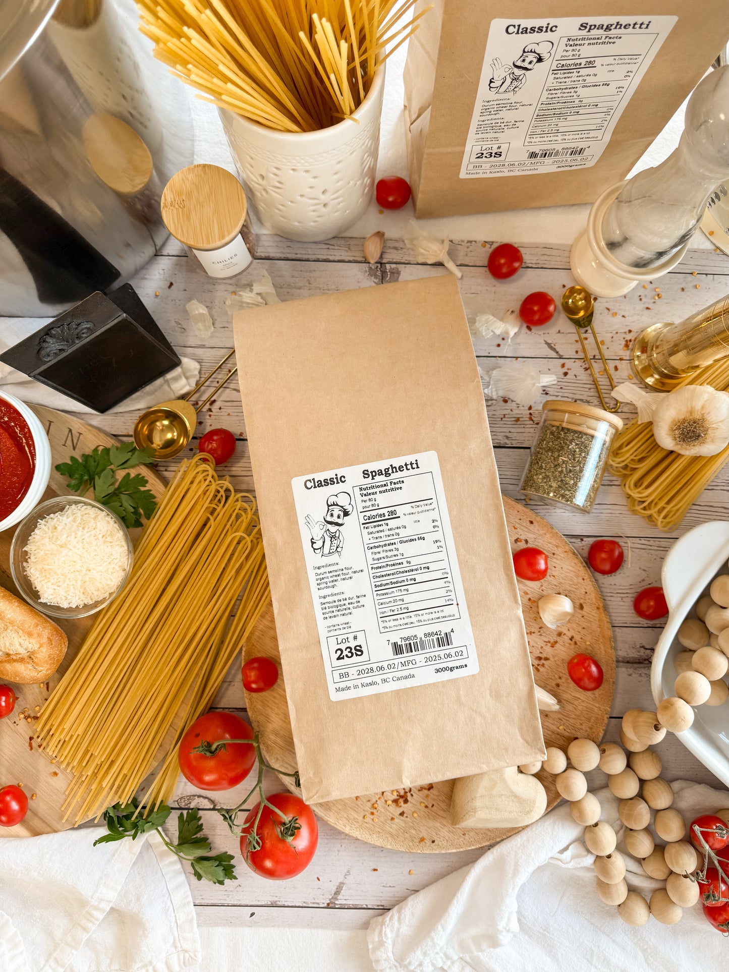 Brown paper bag with Kaslo Sourdough Spaghetti laying on a wooden table with tomatoes and spaghetti around in a lifestyle picture