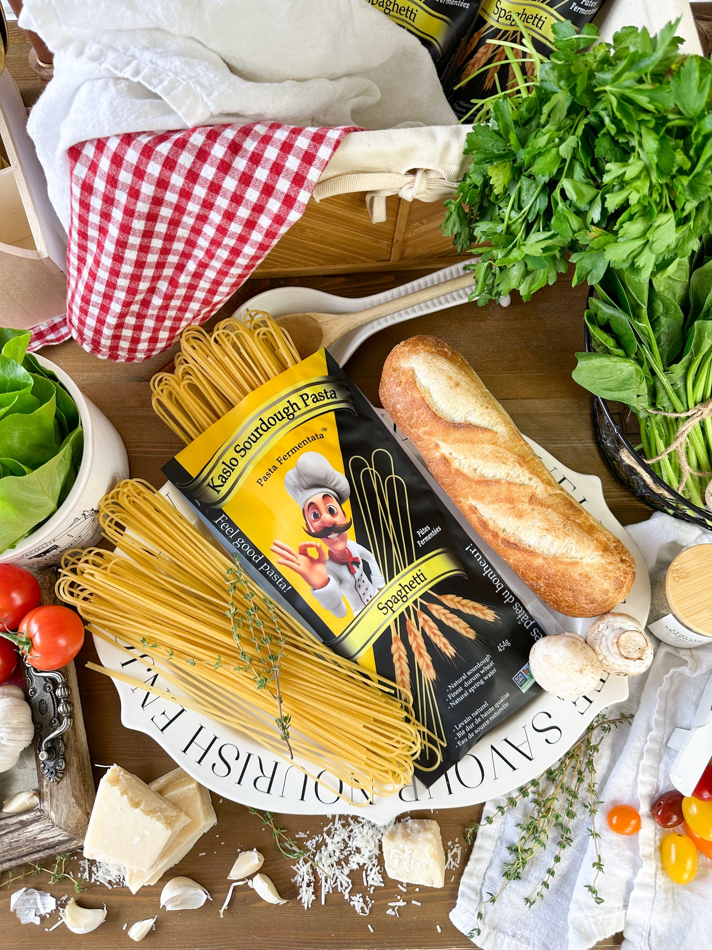 A styled flat-lay of a bag of Kaslo Sourdough Pasta on a white platter with spaghetti, a loaf of bread, garlic, mushrooms, tomatoes, herbs, and baskets on a wooden table.