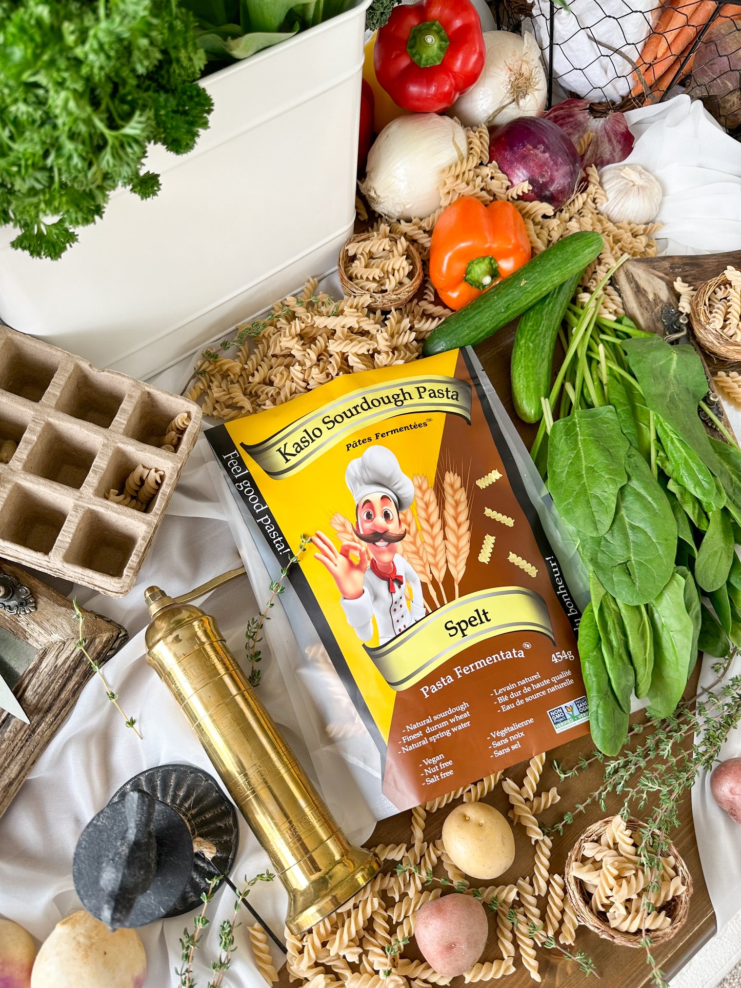 A bag of Kaslo Sourdough Pasta Spelt is styled in a flat lay surrounded by fresh ingredients like spinach, garlic, onions, and tomatoes, along with dried pasta, a brass mortar and pestle, and a seedling tray.
