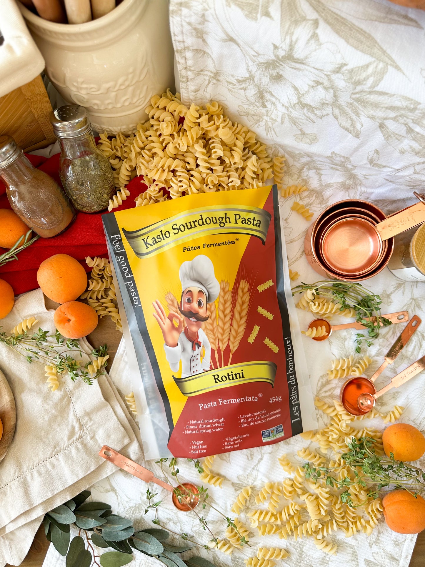 A bag of Kaslo Sourdough Pasta Rotini is displayed in a styled flat-lay scene with raw ingredients and kitchen props, including loose rotini pasta, fresh oranges, copper measuring cups, and herbs on a white patterned tablecloth.
