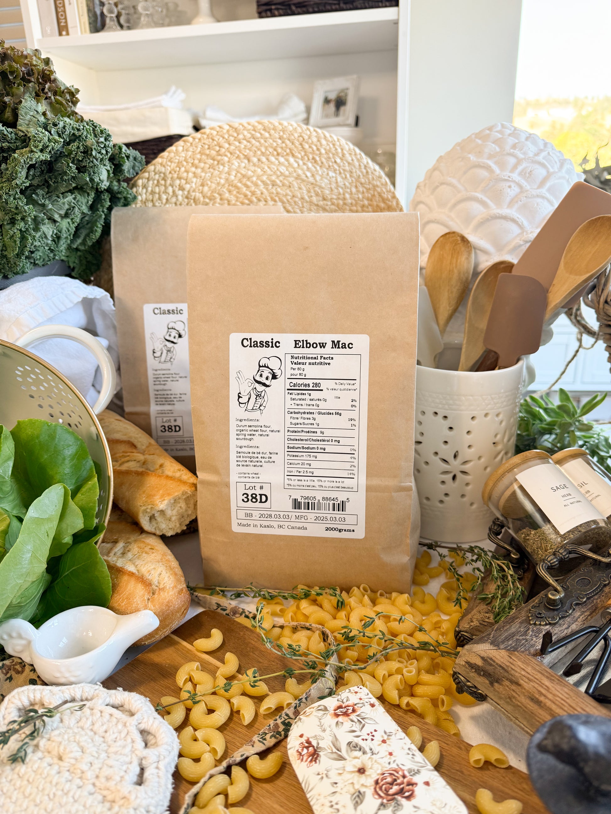 A kraft paper bag of Classic Elbow Mac pasta is displayed on a rustic wooden table surrounded by fresh ingredients, bread, and kitchen utensils.