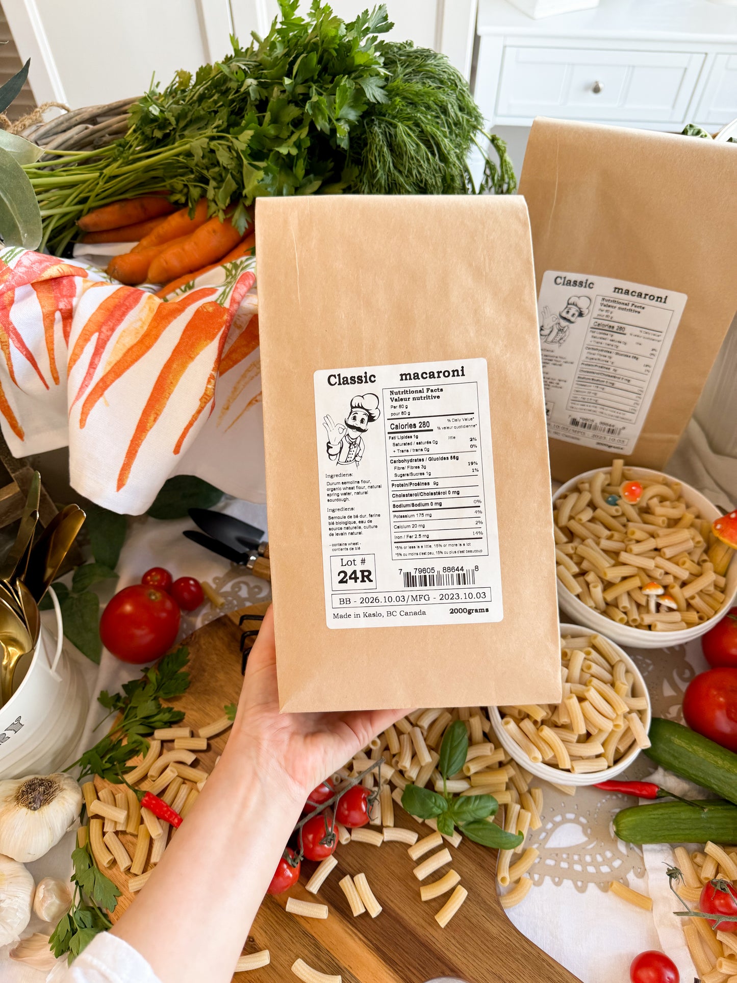A hand holds a brown kraft paper bag of Kaslo Sourdough Macaroni pasta, surrounded by fresh ingredients like carrots, tomatoes, garlic, and basil, with bowls of the prepared pasta in a kitchen setting.