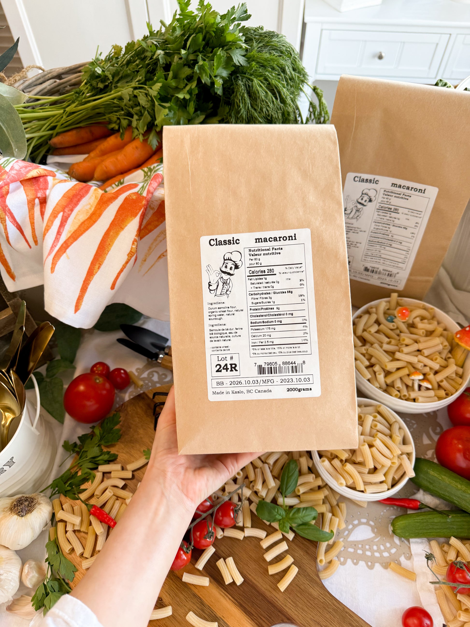 A hand holds a brown kraft paper bag of Kaslo Sourdough Macaroni pasta, surrounded by fresh ingredients like carrots, tomatoes, garlic, and basil, with bowls of the prepared pasta in a kitchen setting.