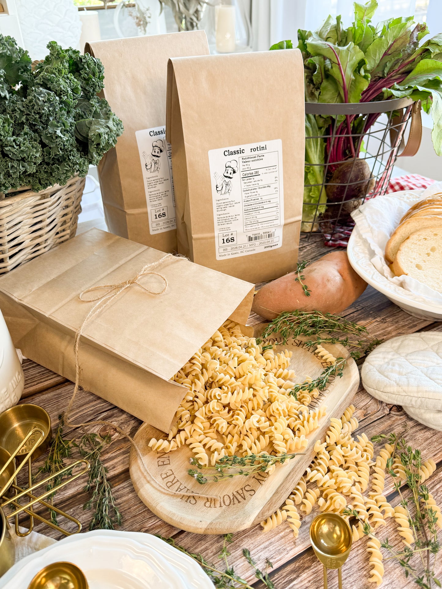 Two brown kraft paper bags of pasta are arranged on a rustic wooden table with fresh vegetables, bread, and gold-toned serving utensils. One bag is open with rotini pasta spilling out onto a wooden board.