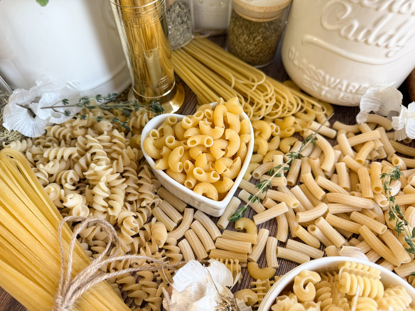 An assortment of uncooked pasta shapes spread out on a wooden surface, including rotini, linguine, Radiatori, and spaghetti, with a heart-shaped white bowl in the center holding elbow macaroni.