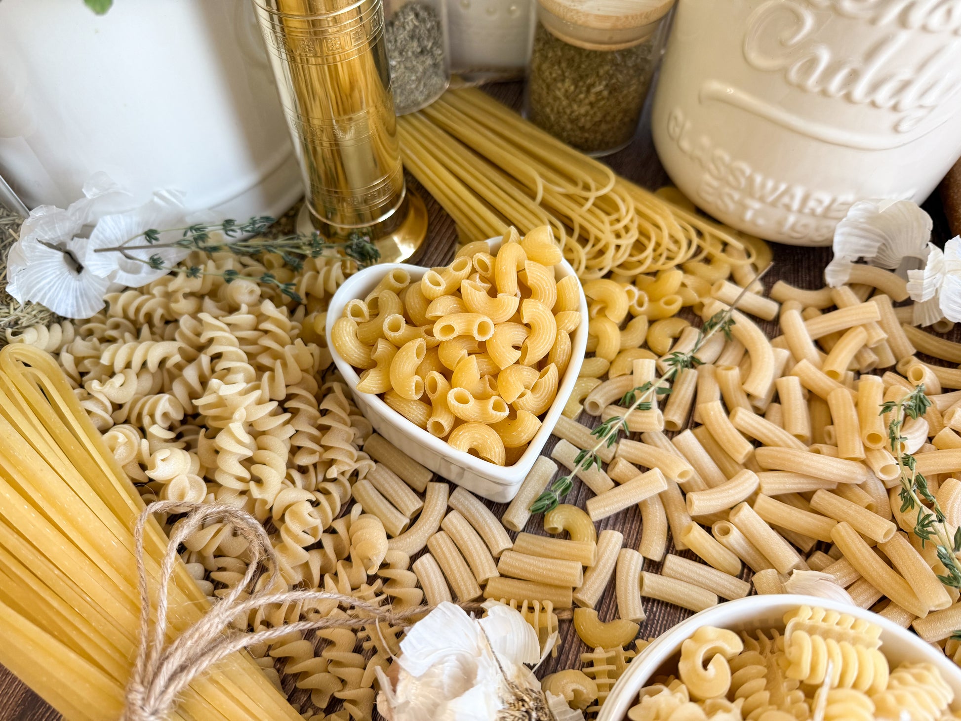 An assortment of uncooked pasta shapes spread out on a wooden surface, including rotini, linguine, Radiatori, and spaghetti, with a heart-shaped white bowl in the center holding elbow macaroni.