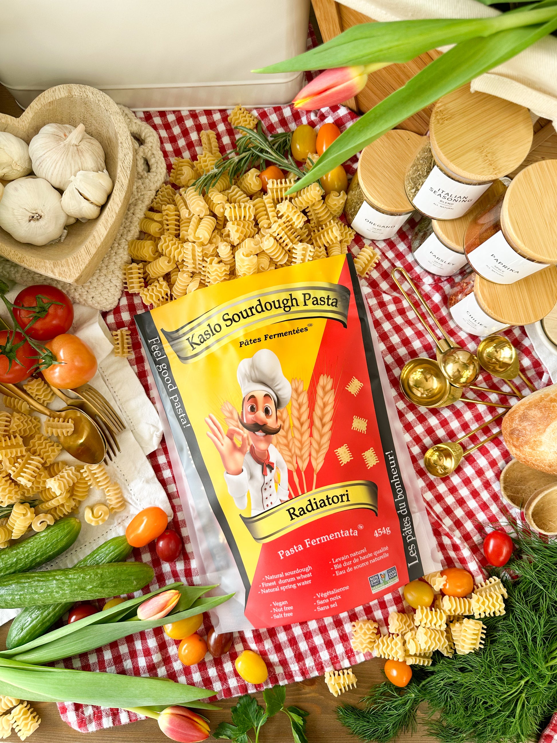 A bag of Kaslo Sourdough Pasta 'Radiatori' is displayed in a beautifully styled flat-lay on a red and white checkered cloth, surrounded by ingredients like garlic, tomatoes, corn, dill, and olive oil jars.