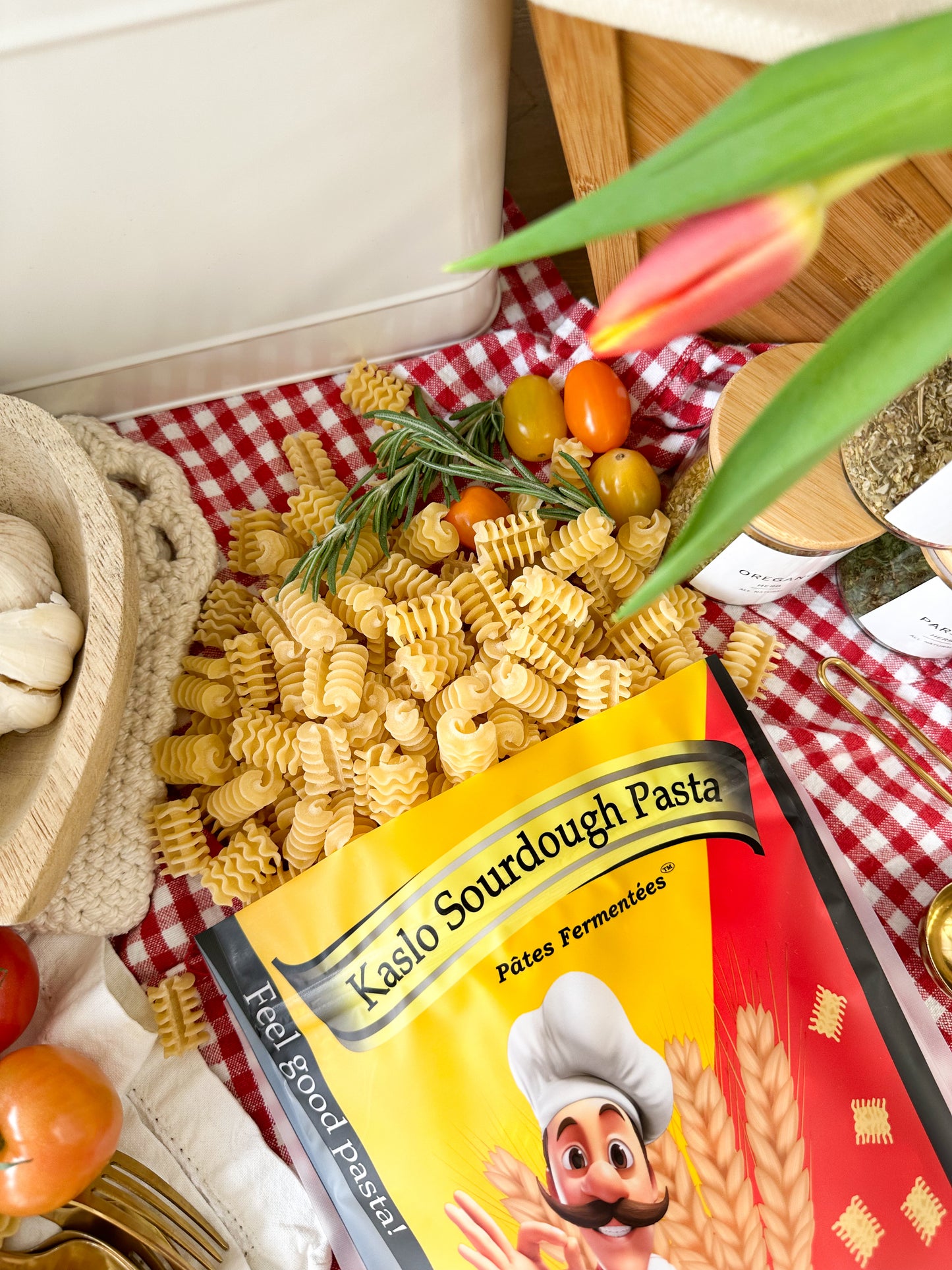 A bag of Kaslo Sourdough Pasta lies on a red-and-white checkered cloth, surrounded by loose radiatori pasta, cherry tomatoes, olives, garlic, herbs, and a tulip leaf in a styled kitchen setting.