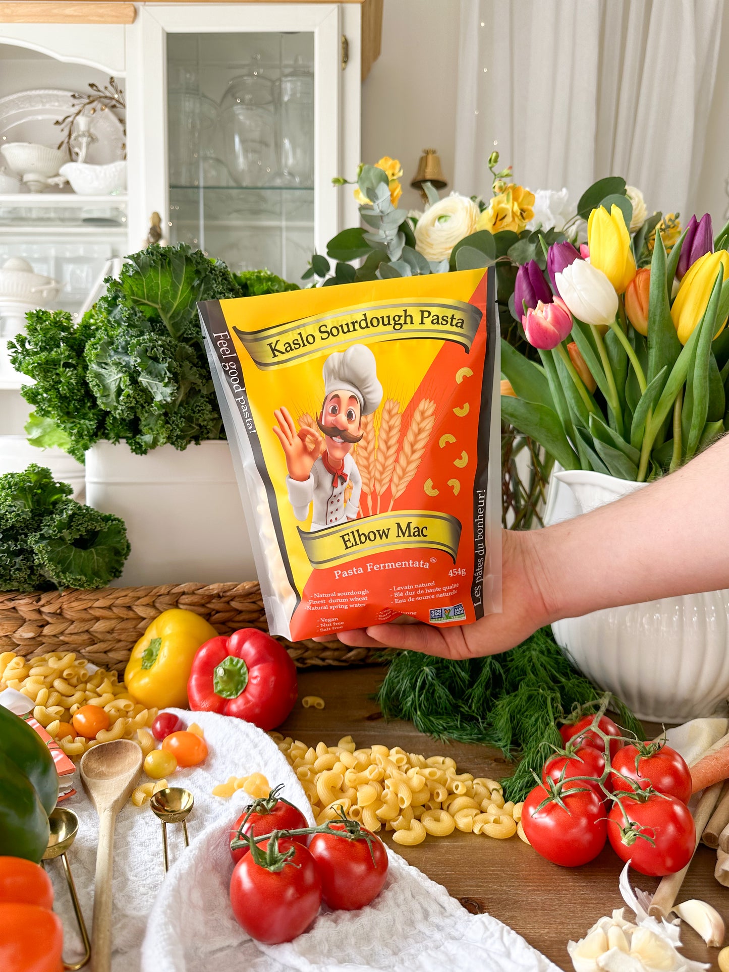 A hand holds a bag of Kaslo Sourdough Pasta Elbow Mac in a kitchen setting surrounded by fresh ingredients like tomatoes, broccoli, garlic, and flowers.