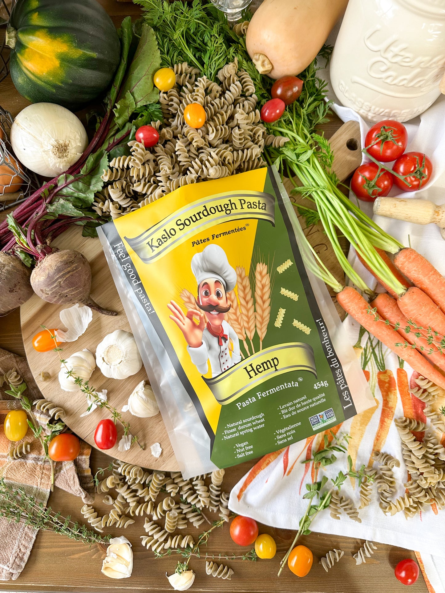 A package of Kaslo Sourdough Pasta Hemp flour is displayed in a styled flat-lay arrangement of whole wheat pasta surrounded by fresh ingredients like carrots, tomatoes, garlic, and herbs on a wooden table.