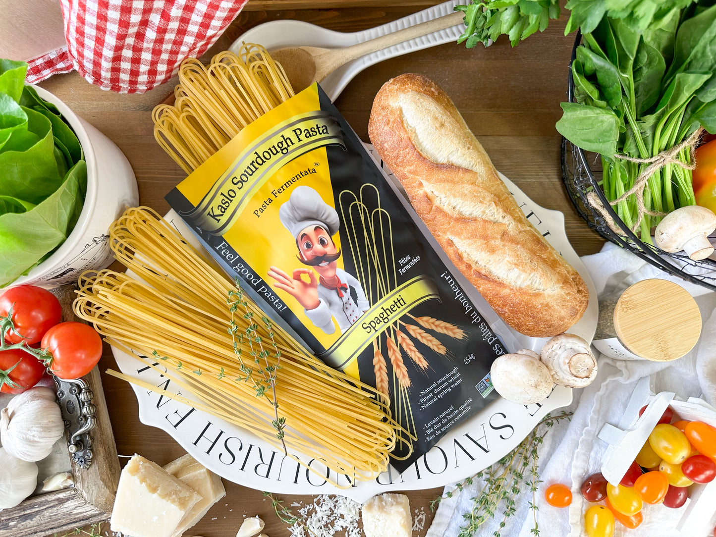 A package of Kaslo Sourdough Pasta Spaghetti lies on a white platter surrounded by baking and cooking ingredients, including a loaf of bread, fresh spinach, tomatoes, garlic, mushrooms, and basil, on a rustic wooden table.