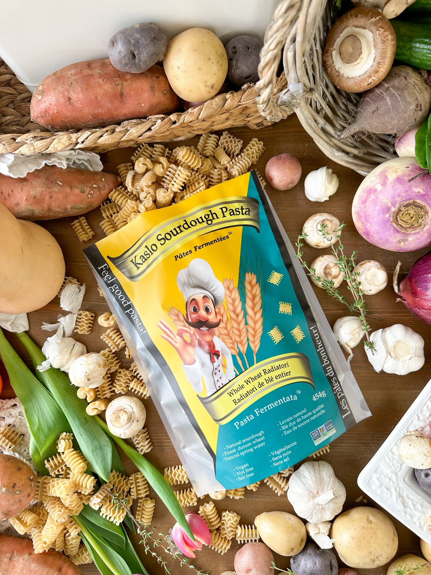A bag of Kaslo Sourdough Pasta is displayed in a styled flat-lay arrangement surrounded by its cooking ingredients, including wheat, garlic, potatoes, onions, and pasta, on a rustic wooden background.