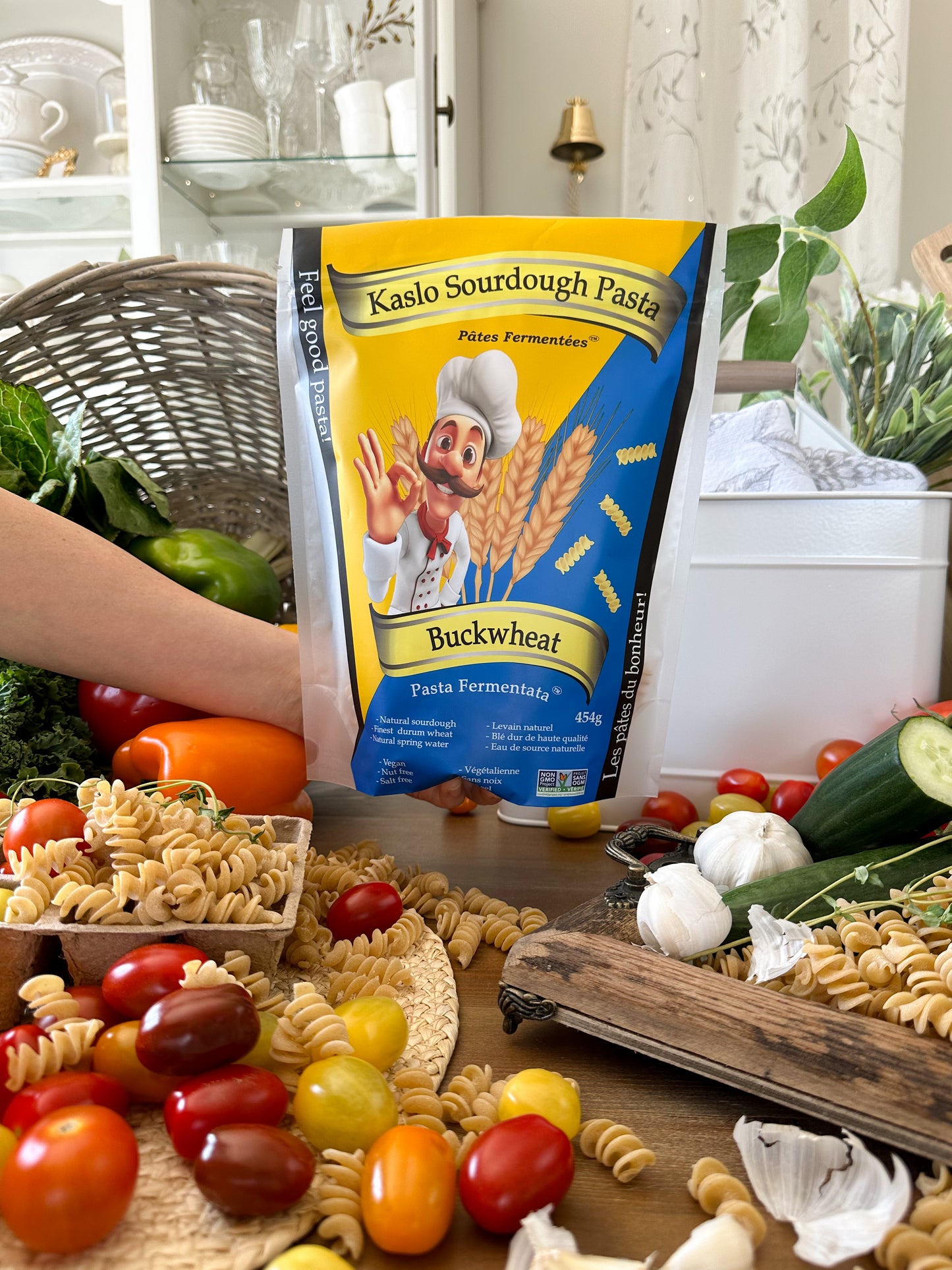 A person's arm is holding a bag of Kaslo Sourdough Pasta Buckwheat against a kitchen backdrop, surrounded by fresh ingredients like tomatoes, garlic, and pasta on a wooden table.