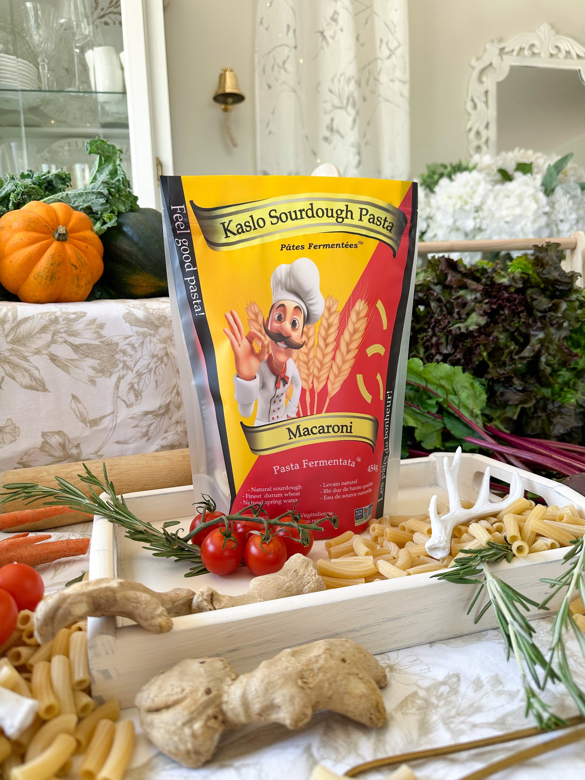 A bag of Kaslo Sourdough Pasta Macaroni stands upright on a styled tabletop with pasta, tomatoes, ginger, rosemary, and vegetables in a kitchen-like setting.