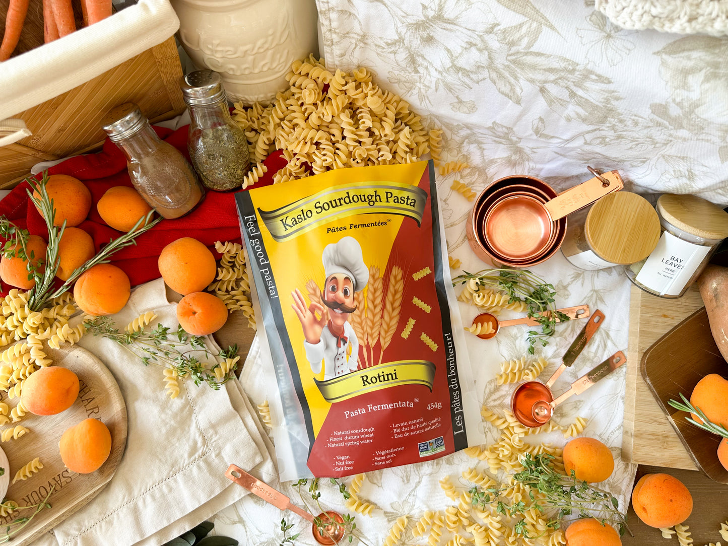 A bag of Kaslo Sourdough Pasta Rotini centered in a styled flat-lay with torn pasta, oranges, measuring cups, spices, and kitchen props on a white patterned cloth.