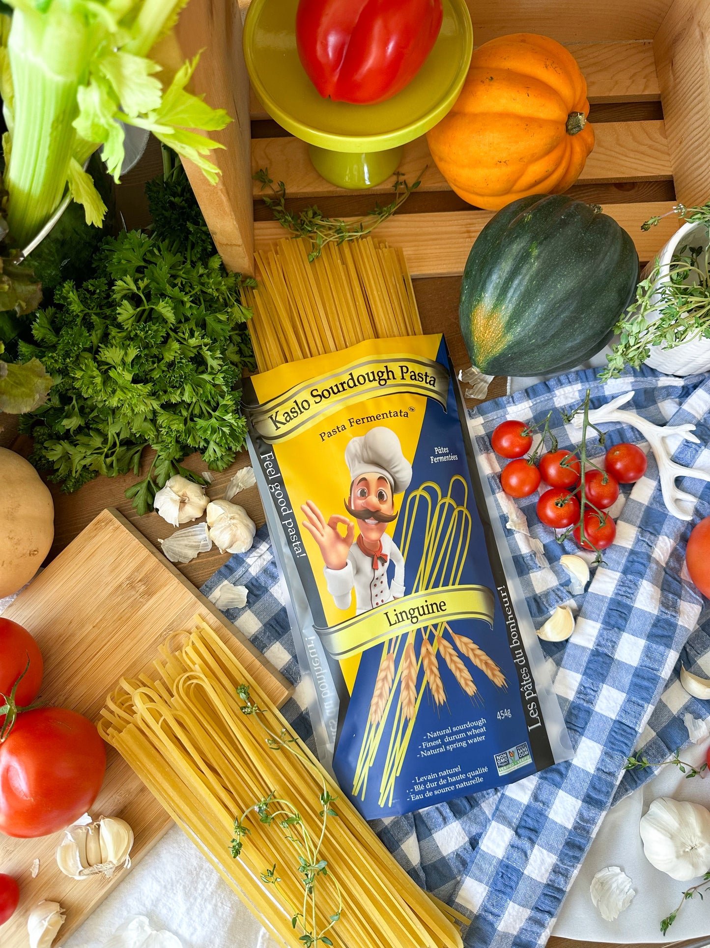 A bag of Kaslo sourdough linguine lays flat on a blue checkered cloth with veggies surrounding the pasta