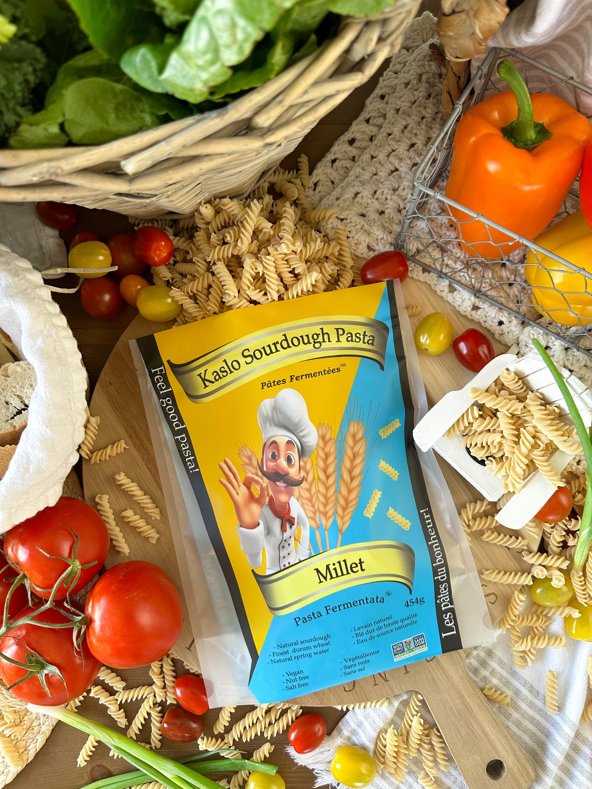 A flat-lay image of Kaslo Sourdough Millet on a wooden cutting board. With pasta and vegetables spread around the background.