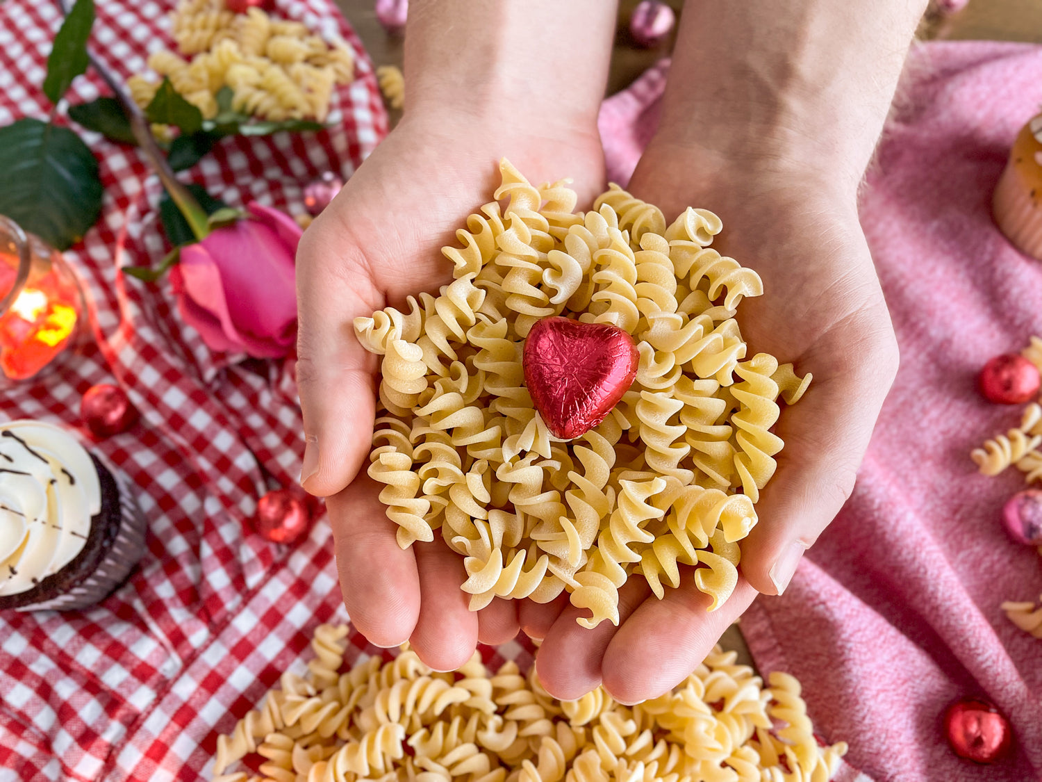 Hands holding a heart-shaped arrangement of pasta with a blurred background of a picnic setup.